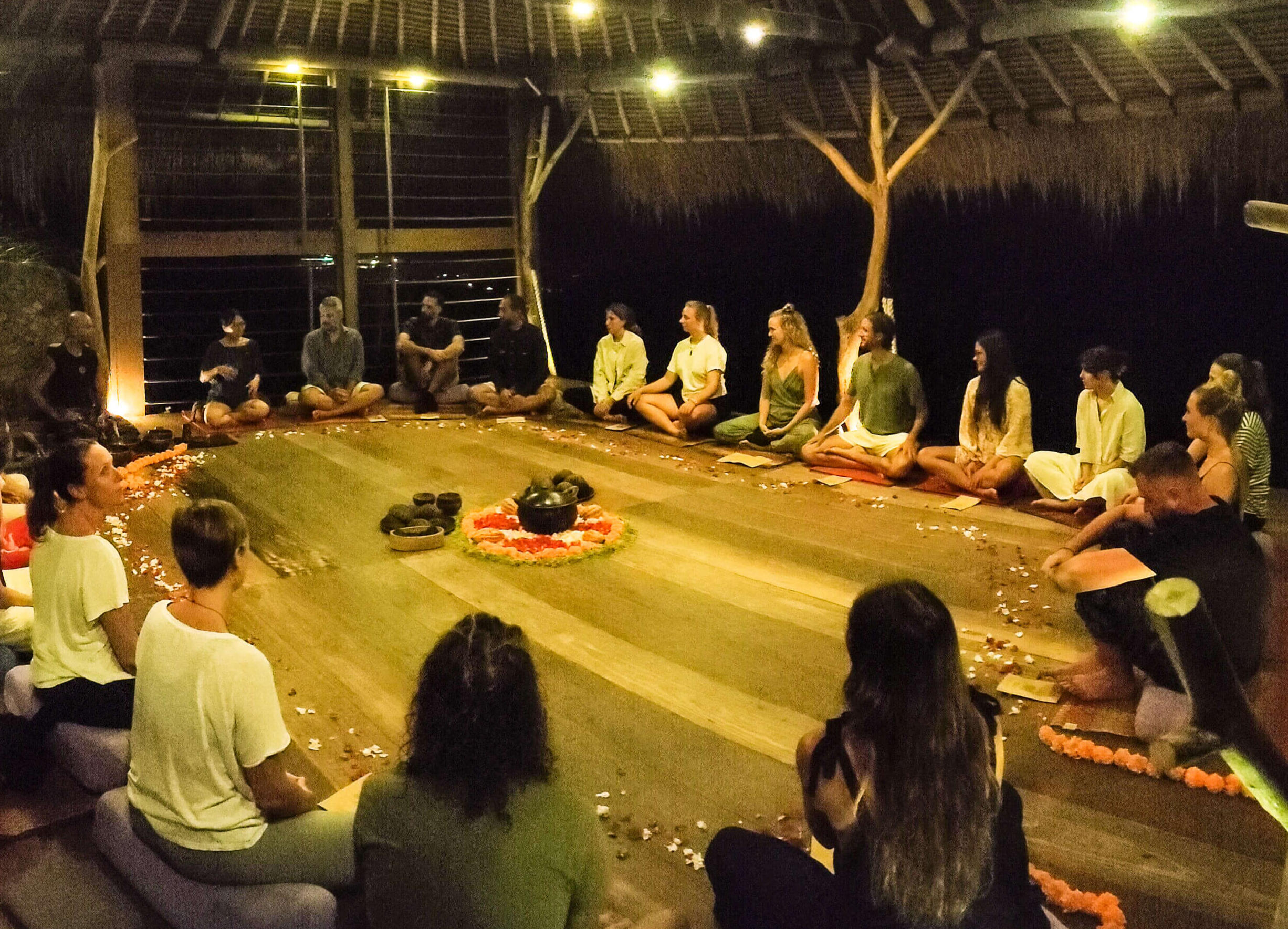 Circle of people gathered in Ashtaris serene yoga shala at dusk, sharing cacao in an intentional ceremony to foster connection, presence, and heart-opening experiences.