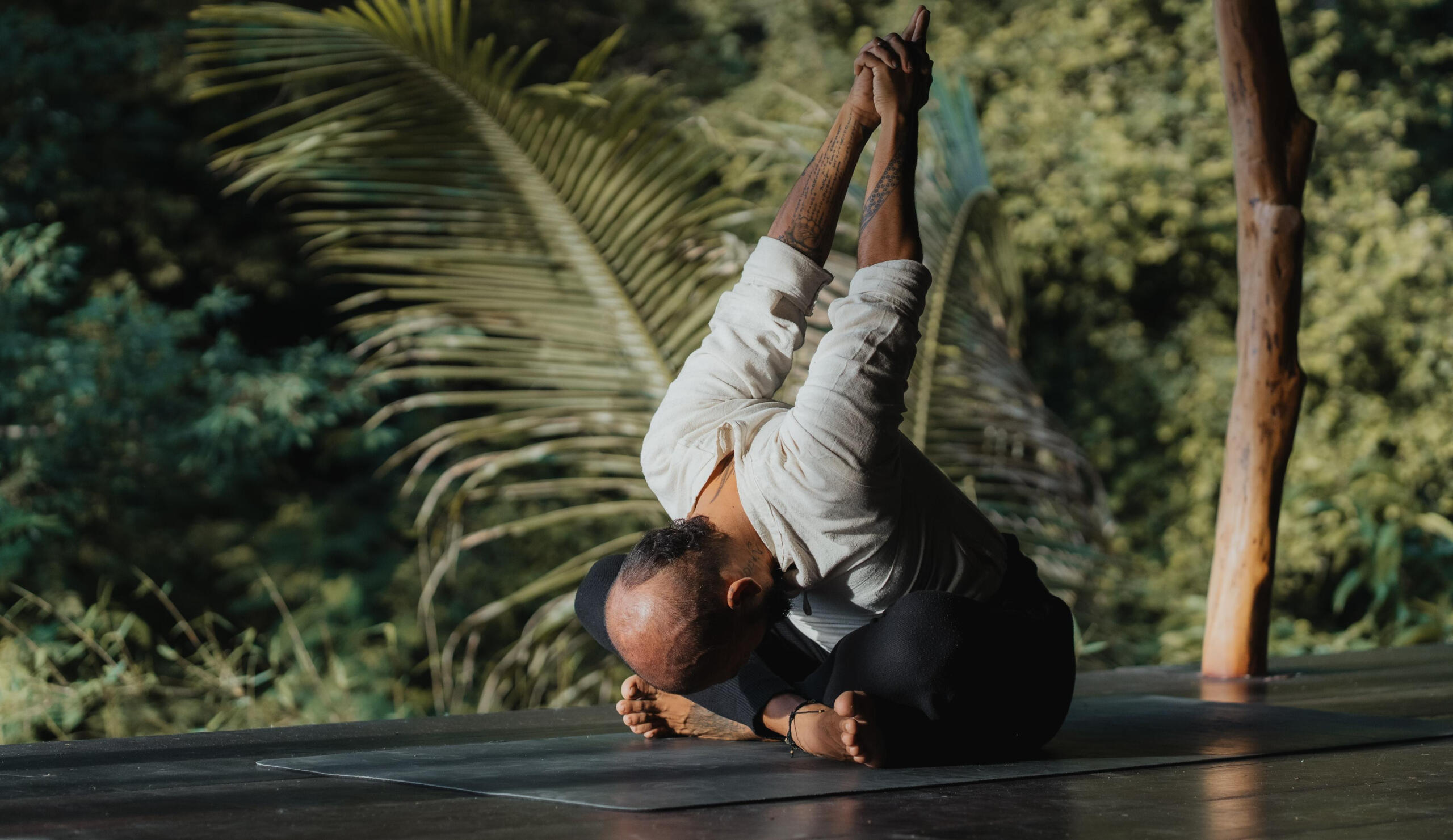 Free Lopez – Seated Forward Fold with Hands Clasped in Lombok in Lush Garden Yoga Shala Free Lopez in his Yoga Practice sitting cross-legged in a yoga shala surrounded by lush tropical greenery in Lombok, folding forward with hands clasped behind his back and lifted toward the sky, expressing openness, surrender, and heart expansion.